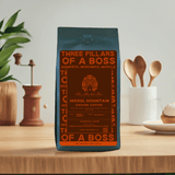 A bag of Mogul Mountain coffee with a label that includes the brand name, the words 'Three Pillars of a Boss', and the scent 'Honesty, Integrity, Might'. The bag is placed on a wooden surface with a plant in the background and kitchen utensils to the side.