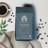 A pack of 'The Mental Bar Treasure Whole Bean Coffee' with a geometric design on the packaging, placed on a linen surface with a cup of coffee and coffee beans scattered around.