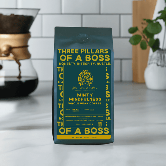 A pack of 'Minty Mindfulness' coffee by The Mental Bar, with the description 'Three Pillars of a Boss - Honesty, Integrity, Hustle'. The pack is placed on a white surface in a home environment, with a plant and a cup in the background.