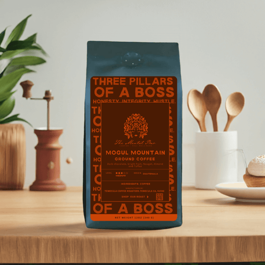 A bag of Mogul Mountain coffee with a label that includes the brand name, the words 'Three Pillars of a Boss', and the scent 'Honesty, Integrity, Might'. The bag is placed on a wooden surface with a plant in the background and kitchen utensils to the side.
